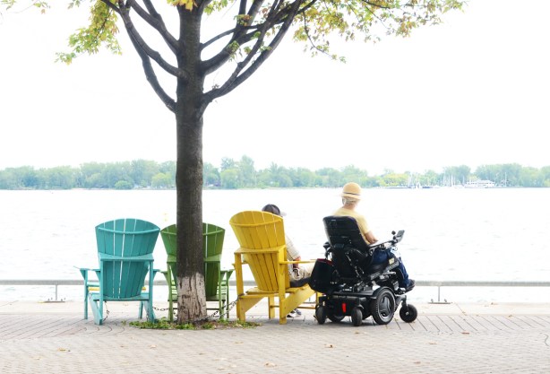 sitting by the waterfront, a man in a yellow Muskoka chair, and a woman in an electric wheelchair, both facing the water
