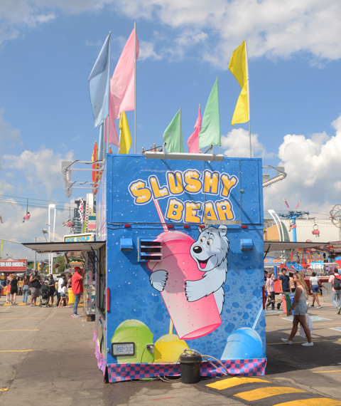 cne midway, a blue wall with a big ad for slushy bear, pastel colour flags on the roof 