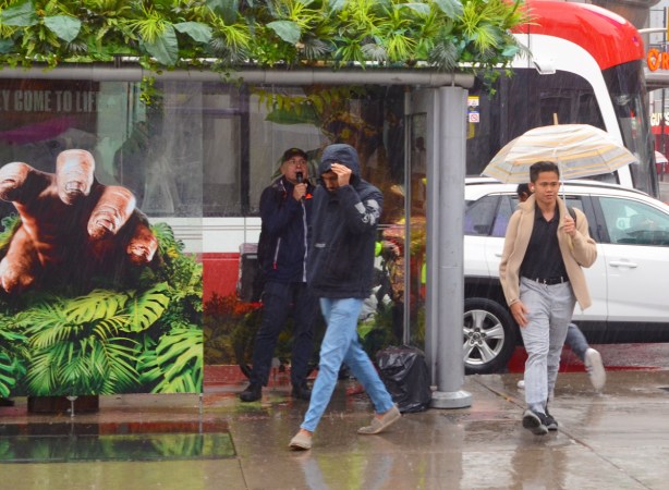 decorated bus shelter, large picture of a hand reaching out, a man in the bus shelter with a microphone, another man walking past with hood up because of the rain, third man with gold and white striped umbrella 
