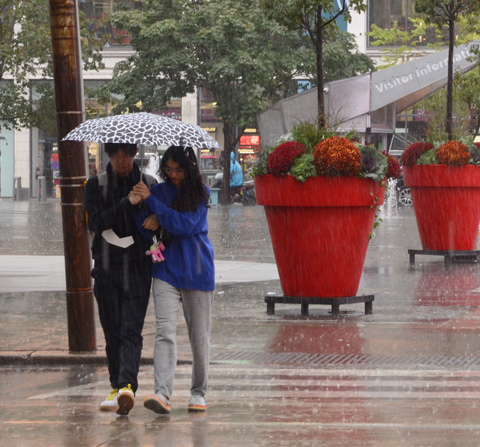 a young couple share an umbrella and walk hand in hand together as they cross Dundas Street in front of the red planters at Yonge Dundas square