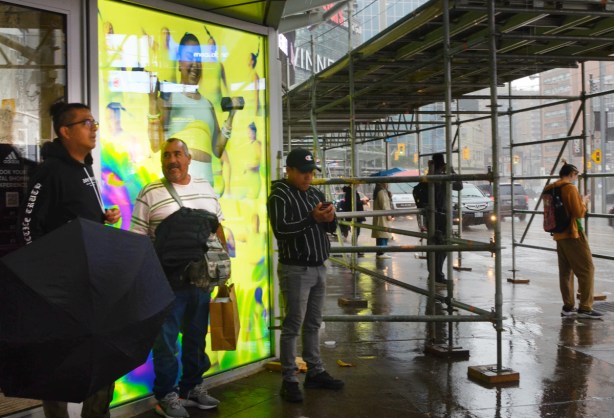 two men talking to each other as they stand beside an electric billboard advert with bright yellow background, other people waiting under scaffolding for the rain to stop