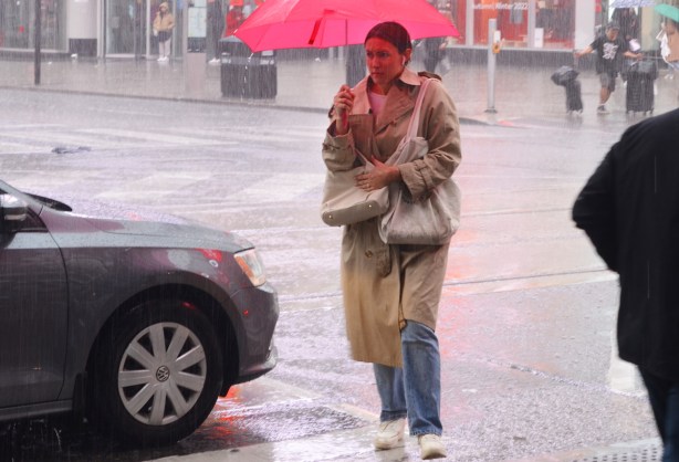 woman with red umbrella and long beige coat walks in front of a car, in the rain, at Yonge and Dundas, 