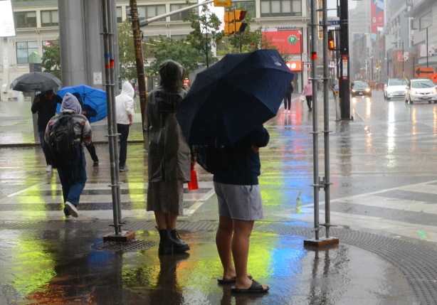 a man in shorts and sandals with a blue umbrella stands beside a woman in black boots and long coat, under shelter while it rains, waiting for a green light to cross Dundas Street 