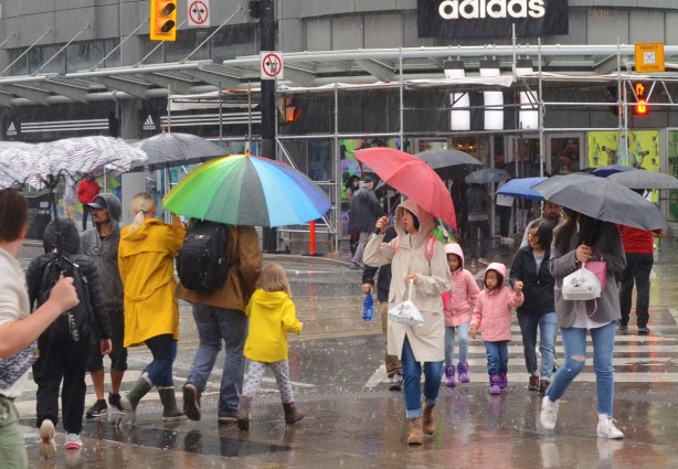people crossing on the diagonal at Yonge and Dundas, kids with pink rain coats, woman with pink umbrella, family with large rainbow umbrella, man with a broken umbrella 