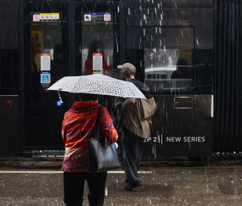 a woman in a red jacket with a white umbrella and black purse, walks towards TTC streetcar doors that haven't opened yet, a man holding up his pants is also waiting for streetcar