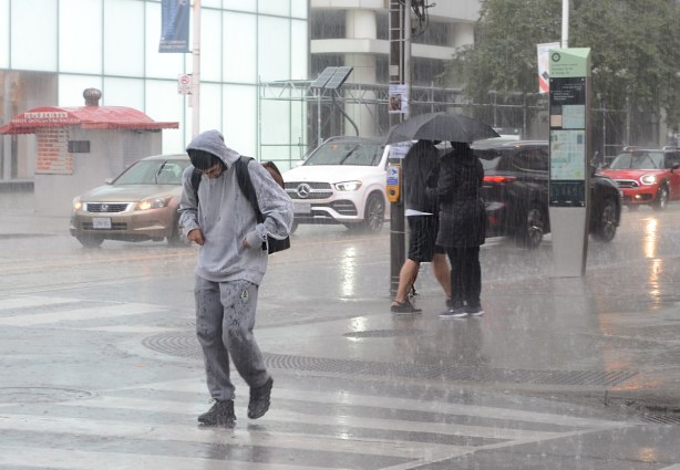 man in grey hoodie walking with head down in the rain as he crosses a wet street 