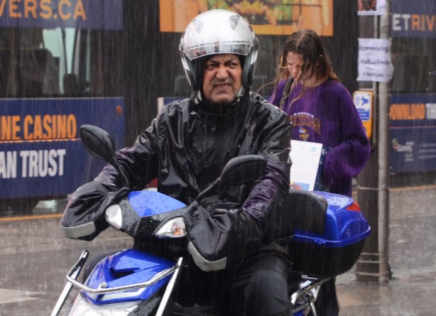 a man on a blue motorbike, in the rain, silver coloured helmet, scowling at the weather, woman behind him has hair blowing in the wind 