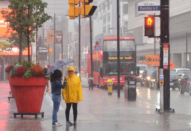 two young women standing in the rain at the corner of Yonge and Dundas, a Hop on hop off tourist bus behind them, large red planters in the square, one woman with a blue umbrella, the other wearing a bright yellow rain jacket with hood up