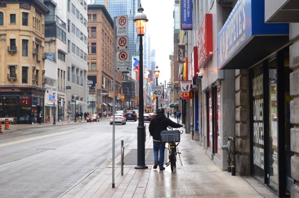 a man walks his bike on the sidewalk, northbound on Yonge street, east side, north of Adelaide