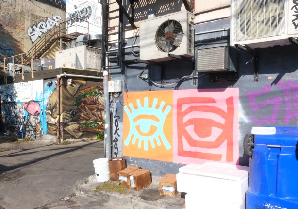 elicser elliott mural in background, 2 abstract minimalistic designs on a wall with air conditioners and other containers, one is red on pink and the other ooks like a green eye and eye lashes on orange paper