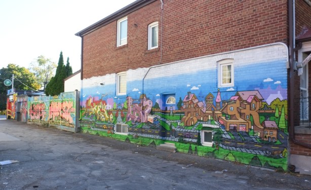 side of a two storey brick house with lower half covered in a mural with stylize street scene, houses, street, cars, green grass, 