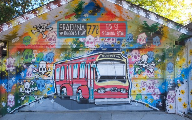 mural by Dudeman covering the front of a garage in an alley, lots of differenc=t coloured skulls, a TTC bus, signs for Spadina