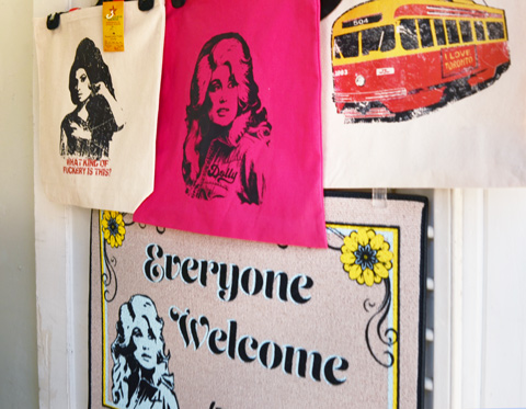 samples of store merchandise on a door, a mat that says everyone welcome, a canvas bag with an image of an old TTC streetcar, a pink bag with a black stenciled womans face on it