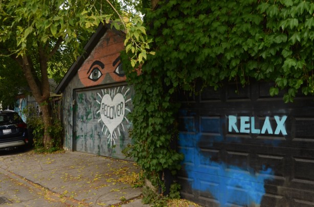 two garages in a lane, ivy covering a lot of the nearest one, the word relax painted on the door, on the second garage, two large eyes above the door, and a white heart on the door with the word recover written in the middle of it 