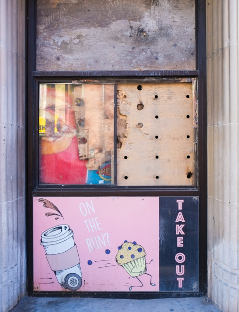 covered windows of closed coffee shop, plywood in one pane, picture of pink and whtie coffee cup and chocolate chip muffin in another pane