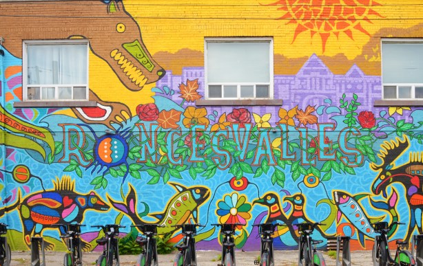 new indigenous theme mural for roncesvalles village, a large brown bear looks down at the text in the mural, a lone of fish and birds under the text, a row of rental bikes in front of the mural, an orange sun in a yellow sky 