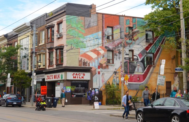 Jac's Milk convenience store on Roncesvalles, with a large mural on the side wall, a TTC streetcar, text that says Roncesvalles village, people waiting for the streetcar