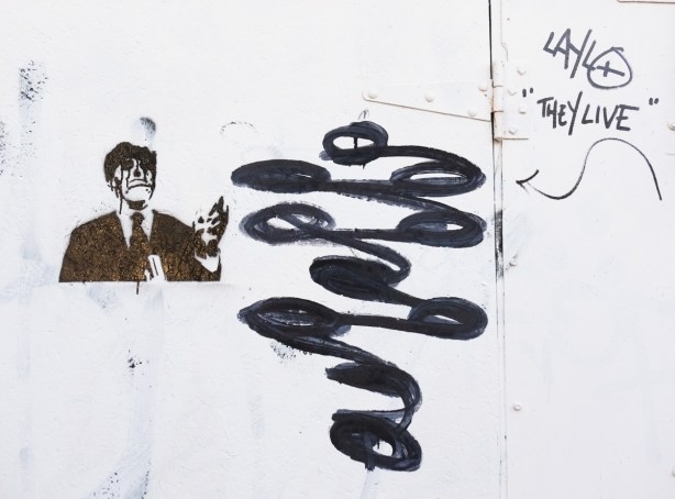 black graffiti on a white garage door, black stencil of man upper body, some blobs and the words they live