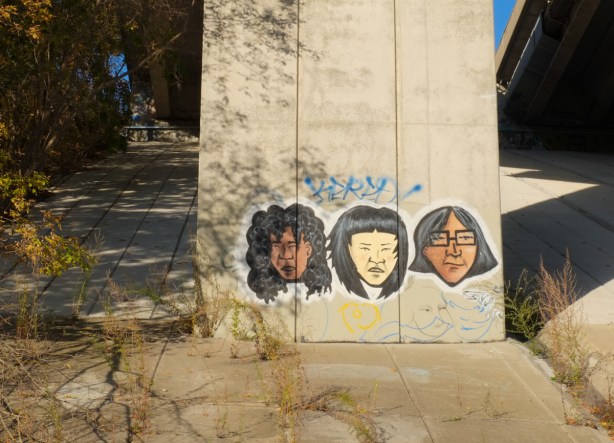 three faces, graffiti on concrete supports for a bridge. each face has black hair, one has glasses and their skin is various shades of brown