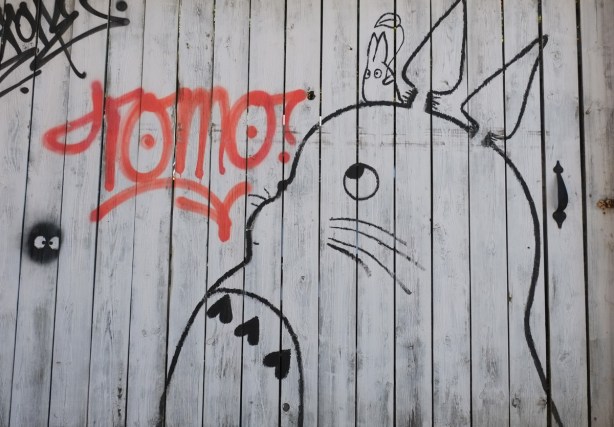 on a wood fence, painted white, with black line drawing of a large plump cat like figure with two ears that stand up, small eyes, whiskers, and 3 hearts on his tummy, a small rabbit sits on its head. red word tomo with question mark beside it