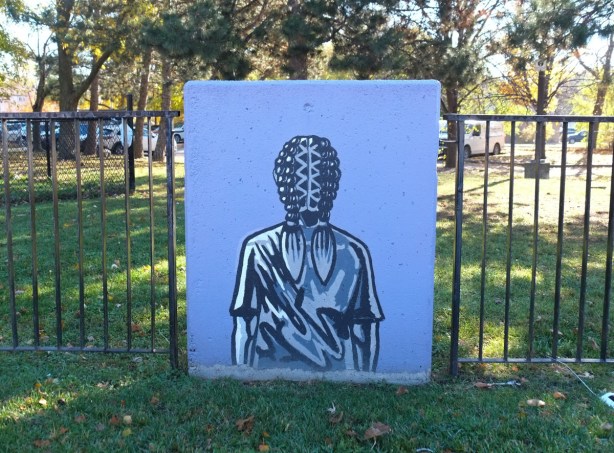 concrete block against a metal fence with simple painting of a person's back, braided hair down middle of head