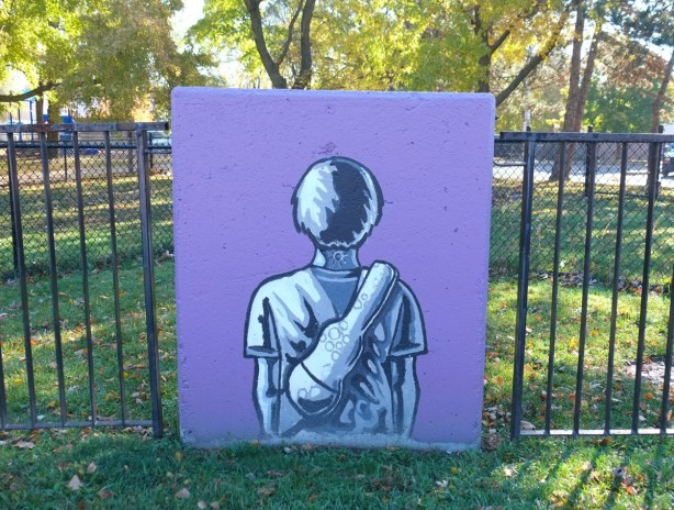 concrete block against a metal fence with simple painting of a person's back, short hair, tattoo of sun on back of neck, carrying a violin case on his back