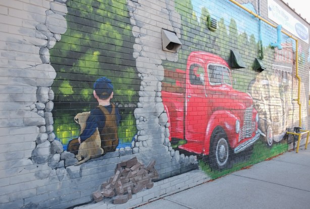mural in Highland Creek, painted grey brick wall, the front of a vintage red truck has come through the wall, pile of bricks beside, a young boy in blue cap and brown overalls sits with his dog in another hole in the wall 