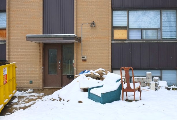 a blue arm chair and a wood chair in the snow beside a snow covered pile of rubble, a yellow bin, in front of a small apartment building 