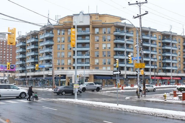 southwest corner of Bathurst and Lawrence, midrose apartment building that curves around the corner, retail at street level, two older people with walkers crossing at the intersection