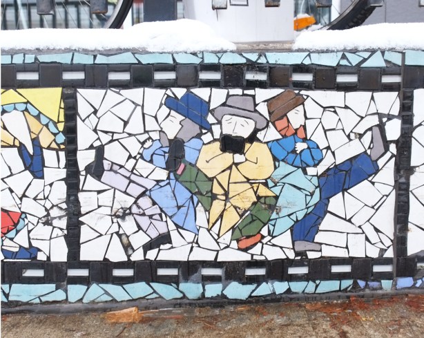 three men dancing, with their backs together supporting each other as they kick their legs out with arms folded 