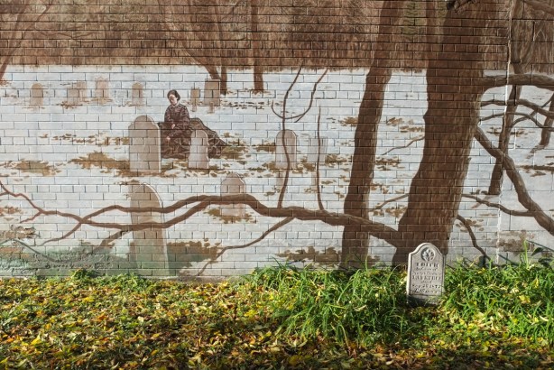 old small tombstone in a cemetery with a wall behind it, mural on wall of a woman sitting by grave stones in a cemetery
