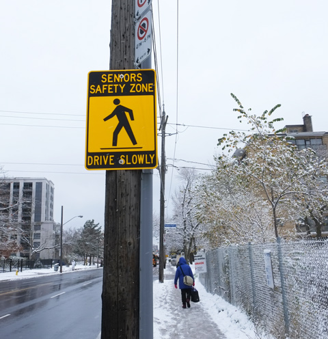 a woman walking up snowy sidewalk on Bathurst, past a pole with a yellow Seniors Safety Zone sign