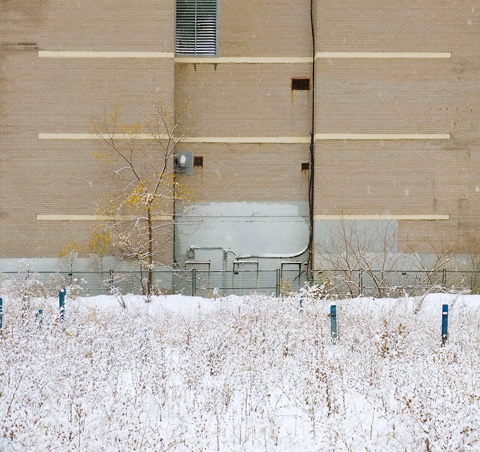vacant snow covered lot in the foreground, black brick wall of apartment building in background, 