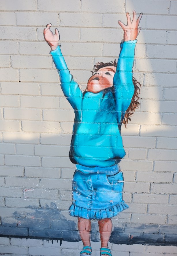 part of a mural, a young girl in blue top and blue shorts, arms upraised, like she is asking to be picked up
