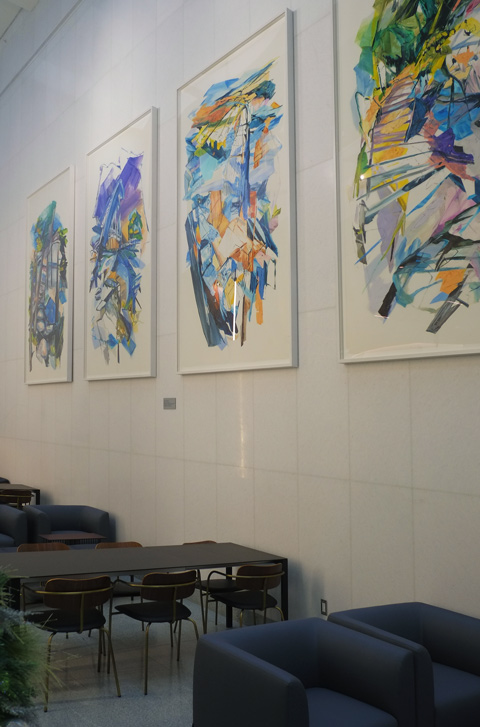 four water themed paintings by Linda Martinello hang on a white wall, above chairs and desks.