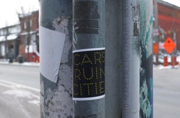 sticker on a metal utility pole on Lansdowne, black with grey text that says cars ruin cities