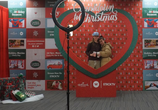 2 young women taking a selfie at a specially set up space with proper lighting, and background designed with red and a large green heart, words say Celebrating Christmas