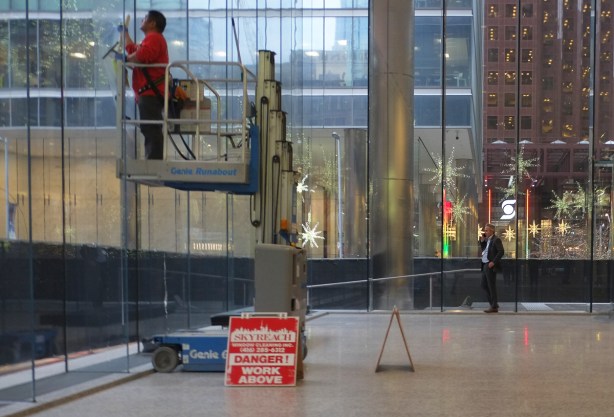 man on a lift washing interior windows, office building lobby, with danger sign below him, man in background looking out window while talking on the phone, christmas lights and decorations outside