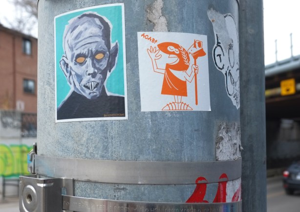 stickers on a pole, one is portrait of man with black gold eyes and large fangs for teeth, the other is a cartoon like drawing of a roman soldier in orange 