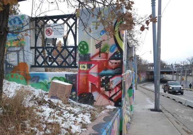 graffiti on concrete supports at the end of a railway underpass, winter time, some snow on the ground, trees, a truck driving past, 
