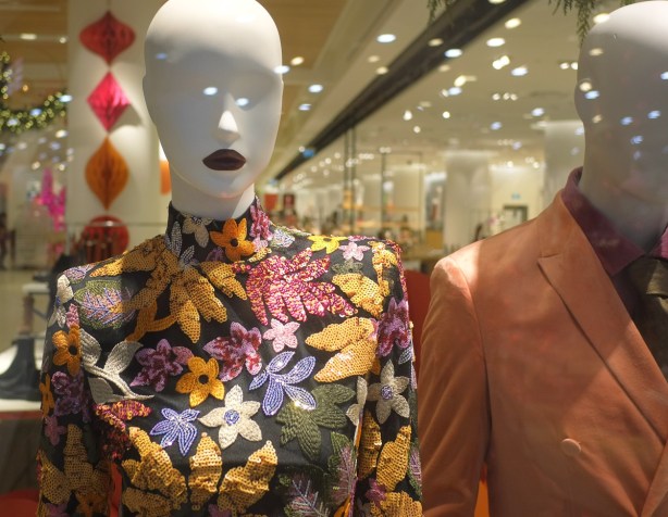 white eyeless mannequin with bright red lipstick, wearing a high necked embroidered top with flowers and leaves on black background, in a store 