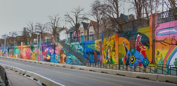 long stretch of concrete wall alongside a railway underpass on Lansdowne Ave., covered with many different murals,