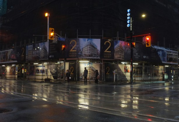 early on a dark and wet morning, the northwest corner of Queen and Yonge, 2 Queen West, a building under renovation and behind hoardings, people waiting on the sidewalk for traffic lights to turn green, reflections on wet pavement
