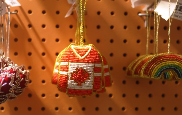 beaded christmas tree ornaments for sale, hanging on hooks, red maple leaf with silver accents on the left, a red and white hockey shirt with maple leaf in center, and a rainbow on the right