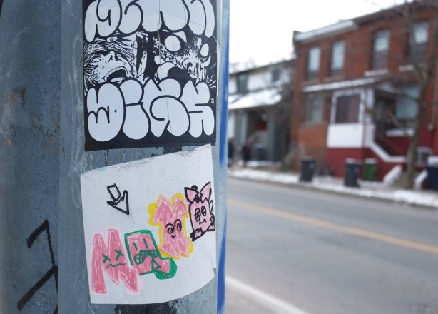 two stickers on a pole, on top is black and white tag like drawing. one on bottom is four little characters in pink with outline and features in darker colours 