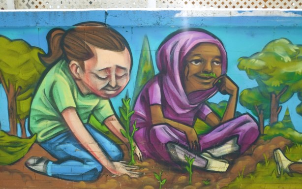 two people sitting in a garden, part of a mural on a wall, one person in a green t shirt is planting a small green twig in the brown dirt, the other person is female and has a purple head scarf