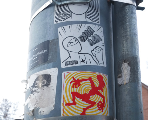 stickers on a metal utility pole, two are urban ninja squadron, and one has a red silhouette on concentric circles of yellow and white