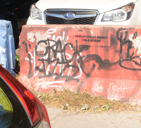 spray painted in black, stenciled text on concrete barrier beside parking lot says revolutionary change for a dollar