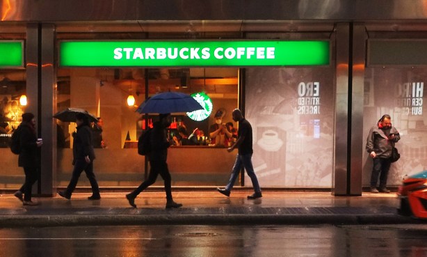 people walking past Starbucks on Yonge Street, morning, still darkish outside, people inside the coffee shop, man with umbrella outside, another man standing by wall looking at his phone
