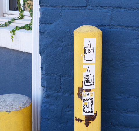 in front of a very blue wall, a yellow bollard with three stickers on it. The phrase let me tell you is written on the three stickers